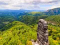 Skalny filar góruje nad zielonymi lasami w Czechach, blisko granicy. Krajobraz pełen jest drzew i skalistych zboczy.
