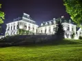 Elegancki hotel w lesie nocą, oświetlony blaskiem okien i zewnętrznych lamp. Otoczony zielenią, zaprasza do odpoczynku.