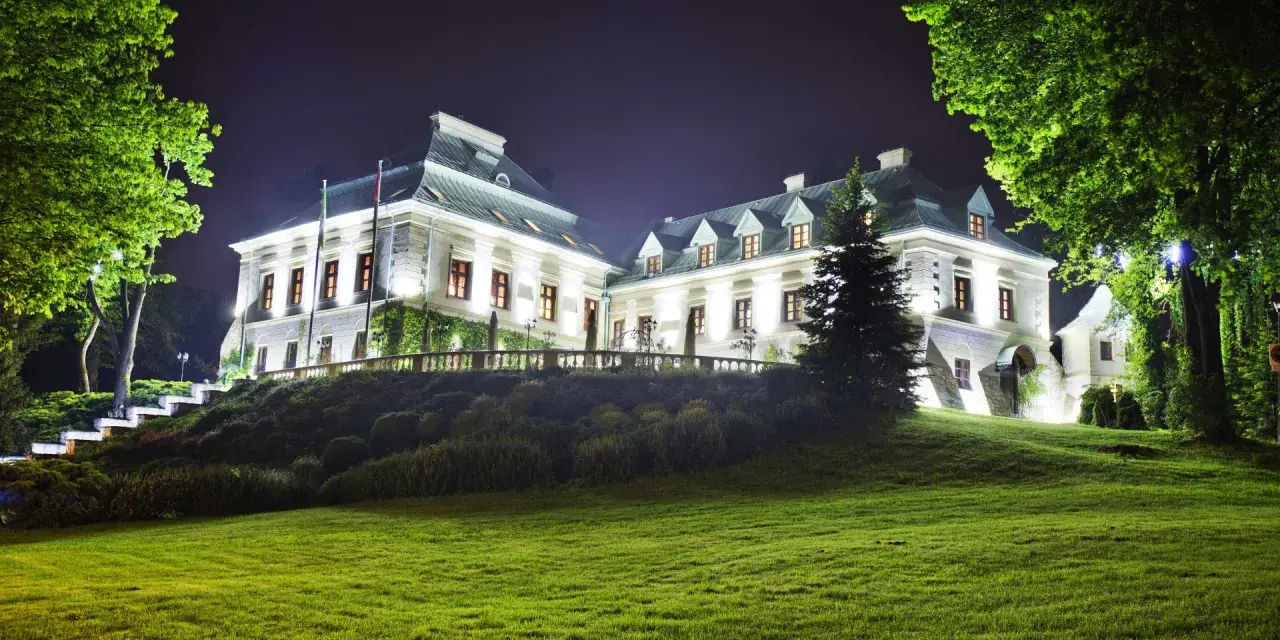 Elegancki hotel w lesie nocą, oświetlony blaskiem okien i zewnętrznych lamp. Otoczony zielenią, zaprasza do odpoczynku.