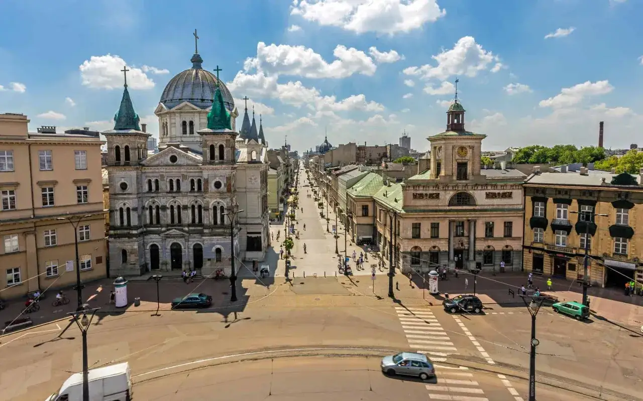 Widok na ulicę Piotrowską w Łodzi, z kościołem św. Mateusza i kamienicami. Spacerowicze i samochody na tle błękitnego nieba.